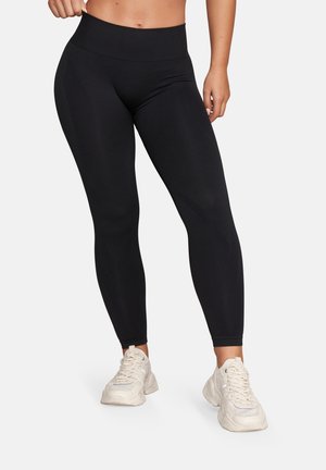 Kobieta w czarnych legginsach z wysokim stanem i masywnych białych trampkach, stojąca na tle jednolitego białego tła.