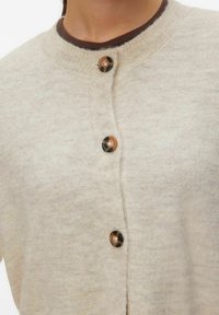 Bež pletena jopica z vrhovnim izrezom in petimi zaobljenimi gumbi v dveh tonih. Mehka tekstura in subtilen pikčast vzorec po celotni površini.