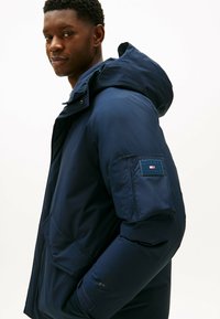 Donkerblauwe gewatteerde jas met een capuchon, voorzien van een logo-patch op de linkermouw en zakken aan de voorkant. Gemaakt van glad, weerbestendig materiaal.