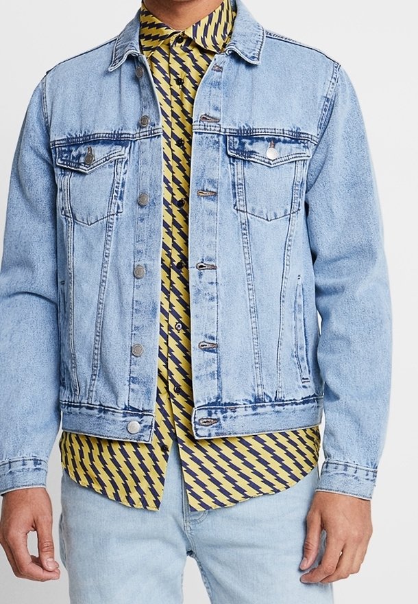 Homme portant une veste en denim bleu clair sur une chemise jaune avec un motif d'éclairs noirs, associé à un jean bleu clair.