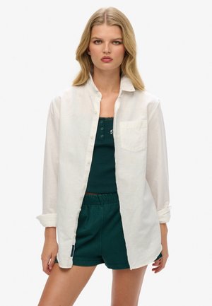 Biała, długorękawowa, zapinana na guziki koszula wykonana z lekkiego materiału, nałożona na ciemnozielony top na ramiączkach i szorty. Posiada kieszeń na piersi i podwinięte mankiety.