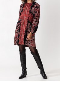 Schwarzes und rotes Paisley-Muster-Kleid mit langen Ärmeln und schwingendem Rock, kombiniert mit knielangen schwarzen Lederstiefeln.