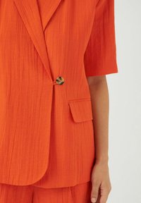 Oranssi blazer, jossa on teksturoitu kangas, lyhyet hihat, yksittäinen nappisulku ja paikkatasku. Näkyvät ompeleet lisäävät yksityiskohtia.