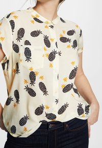Blouse à manches courtes avec boutons, couleur crème, ornée d'un motif d'ananas noir avec des accents jaunes et une seule poche poitrine. Texture lisse.