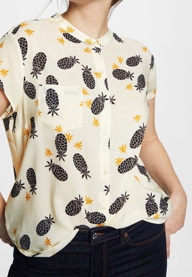 Blouse à manches courtes avec boutons, couleur crème, ornée d'un motif d'ananas noir avec des accents jaunes et une seule poche poitrine. Texture lisse.