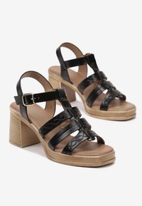 Sandales ouvertes en cuir noir avec des lanières épaisses tressées, un talon carré et une semelle texturée. Bride de cheville ajustable avec détail de boucle.