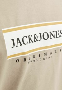 Beige T-Shirt aus weicher Baumwolle. Mit einem auffälligen weißen und goldenen Grafikdesign sowie dem Text "JACK & JONES ORIGINALS WORLDWIDE".