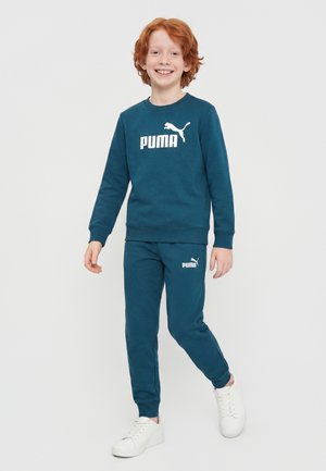 Rødhåret barn smiler, iført en teal Puma sweatshirt og matchende bukser, går med hvide sneakers på en ensfarvet lys baggrund.