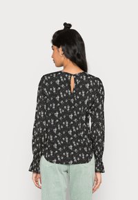 Blusa preta com padrão floral, featuring mangas longas, um detalhe de abertura nas costas em forma de chave e uma ligeira curva na bainha. O material parece leve e com um acabamento texturizado.
