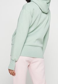 Felpa verde menta con tasca a marsupio e orlo a costine. Presenta una consistenza morbida e una vestibilità comoda, abbinata a pantaloni rosa chiaro.
