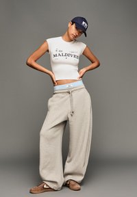 Hvid cropped t-shirt med "MALDIVES" print, lysegrå sweatpants med snøre og blå talje, brune sneakers. Marineblå kasket med NYC-logo.