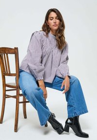 Femme aux longs cheveux bruns assise sur une chaise en bois, portant un blouse à manches bouffantes rayée, un jean bleu et des bottines noires à talons.