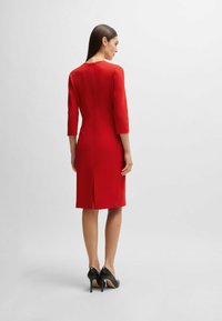 Rotes, tailliertes Kleid mit drei Viertelärmeln, Reißverschluss auf der Rückseite und einem Schlitz am Saum. Getragen mit schwarzen hohen Schuhen.