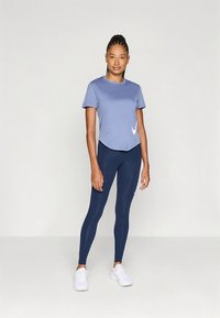 Svetlo modra športna majica s kratkimi rokavi in belim logotipom, kombinirana s temno modrimi pajkicami. Model nosi bele superge in stoji na nevtralnem ozadju.