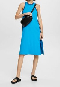 Femme portant une robe midi bleue sans manches avec une fente latérale, un sac bandoulière noir, des sandales noires et des bijoux simples, debout sur fond blanc.