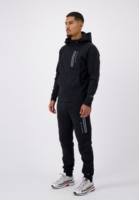 Zwarte zip-up hoodie en bijpassende sweatpants in een zachte stof, met reflecterende accenten en een getailleerd ontwerp, gecombineerd met meerkleurige sneakers.