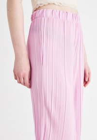 Rozā bikses ar krokām un elastīgu jostasvietu, ar vieglu, gludu tekstūru un vertikālām svītrām, attēlotas jostasvietas līmenī.