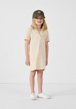 Beige korte mouwen jurk met een V-hals ontwerp, gemaakt van zachte stof. Draagt een donkergroene pet met een embleem en witte sneakers.