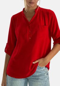Camisa roja de botones con un acabado texturizado, que incluye un bolsillo en el pecho, mangas enrollables y un dobladillo curvado. Fabricada con tela ligera.