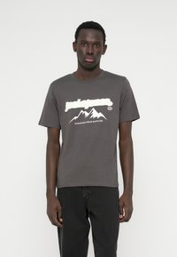 Pelēks kokvilnas t-krekls ar baltu kalnu grafiku un "jackjones" tekstu. Īsas piedurknes un apaļa kakla izgriezums. Apvienots ar melnām džinsu biksēm.