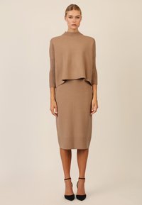 Braunes Strickkleid mit gefaltetem Ausschnitt und drei Viertelärmeln. Der Rock ist figurbetont und wird mit schwarzen High Heels kombiniert.