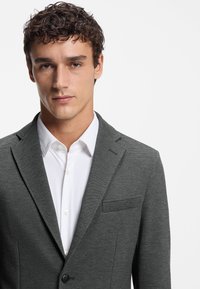 Grey textured blazer with a notched lapel and single button closure, worn over a white collared shirt. Features a chest pocket.