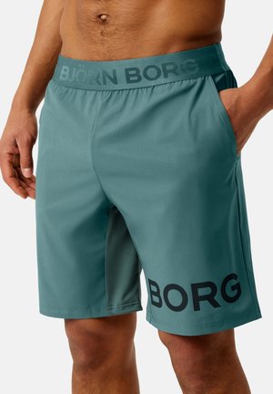 Hombre vistiendo pantalones cortos Björn Borg color verde azulado con cintura elástica y bolsillos laterales, con la mano en el bolsillo derecho, de pie contra un fondo liso.