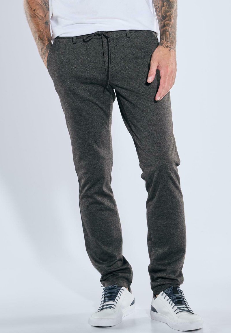Mann trägt dunkelgraue Slim-Fit-Hosen mit Kordelzugbund, weiße Sneakers und zeigt tätowierte Arme vor einem schlichten Hintergrund.