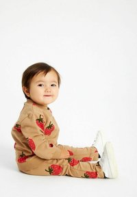 La felpa e i pantaloni jogging color sabbia presentano stampe di fragole rosse. L'outfit ha una vestibilità rilassata, polsini a coste e viene abbinato a sneakers bianche.