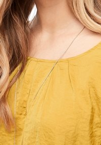 Gul, texturerad blus med samlad detalj vid halsringningen, bärs med ett långt, smalt silversmycke. Håret är vågigt och faller ner förbi axlarna.