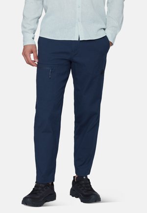 Uomo che indossa pantaloni blu navy con tasca con zip sulla coscia, camicia chiara con bottoni e scarpe da trekking nere, in piedi con le mani nelle tasche.