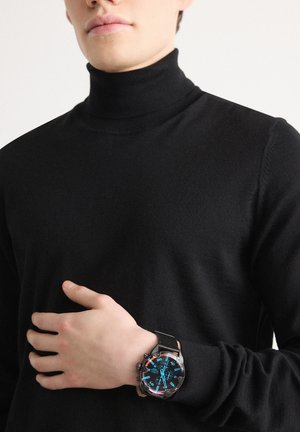 Person, der bærer en sort rullekrave og en sort armbåndsur med en stor blå og rød urskive på venstre håndled.