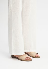 Beige nahkaliukumäet kengät avoimella varpaalla ja ristikkäisellä nauhadesignilla, yhdistettyinä valkoisiin teksturoituihin leveälahkeisiin housuihin.