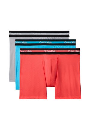 Muskelöser Mann steht barfuß vor grauem Hintergrund, trägt hellgraue Boxer-Shorts mit schwarzem Bund, Arme vor der Brust verschränkt.