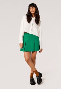 Weißes, kurzes Hemd mit langen Ärmeln und einer einzelnen Tasche, kombiniert mit einem grünen, plissierten Rock. Das Model trägt braune Rippensocken und schwarze Schuhe.