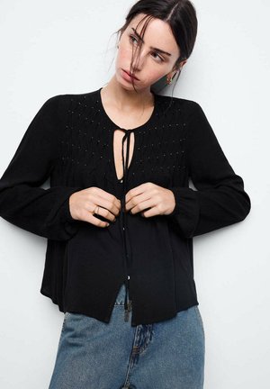 Mujer con blusa negra de manga larga con textura en la parte superior delantera y jeans azules, ajustando los lazos delanteros, el cabello cubriéndole parcialmente la cara.