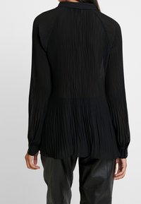 Blusa nera plissettata con maniche lunghe, tessuto morbido e polsini con bottoni; presenta una scollatura alta sulla parte posteriore e una silhouette fluida.