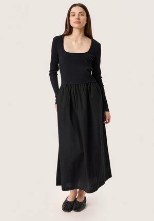 Sort kjole med ribbet overdel, firkantet udskæring og samlet skirt. Lange ærmer og blødt stof. Sorte flade sko med ruchering.