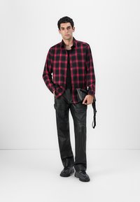 Camisa de cuadros roja y negra de botones sobre una camiseta negra, combinada con pantalones de cuero negros y zapatos negros, sosteniendo una pequeña bolsa negra.
