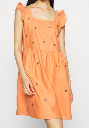 Ærmeløs orange kjole med flæsekantede skulderstropper og små broderede flerfarvede blomster, båret af en stående kvinde.