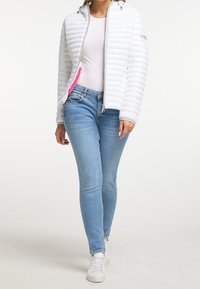 Kobieta w białej lekkiej pikowanej kurtce, białej koszuli, jasnoniebieskich obcisłych jeansach i białych trampkach, stojąca na tle jednolitego tła.