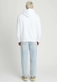 Sweat-shirt à capuche blanc avec texture à côtes verticales, associé à un jean bleu clair et des baskets beiges, vu de l'arrière.