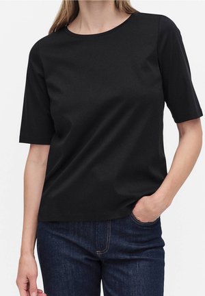Femme portant un t-shirt noir uni à manches courtes et un jean bleu foncé, une main dans la poche, sur fond blanc.