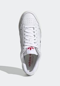 Sneaker bianca con parte superiore in pelle testurizzata, punta tonda e lacci bianchi. Presenta un marchio rosso sulla linguetta e accenti neri sui lati.