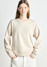 Beżowy sweatshirt z teksturowanym, haftowanym wzorem na środku. Okrągły dekolt i opuszczone ramiona; miękka, gładka tkanina.