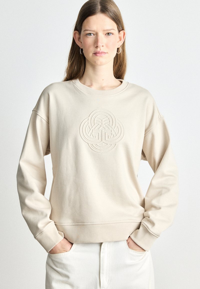 Beżowy sweatshirt z teksturowanym, haftowanym wzorem na środku. Okrągły dekolt i opuszczone ramiona; miękka, gładka tkanina.