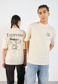 Béžové tričko s krátkými rukávy. Na předn�í straně je nápis "Expresso Club" hnědým písmem; na zadní straně je obrázek cartoonového hrnku kávy a nápis "Expresso."
