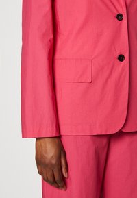 Blazer rose en tissu lisse et léger, doté d'un seul bouton noir, d'une poche latérale et d'une manche à coupe droite.