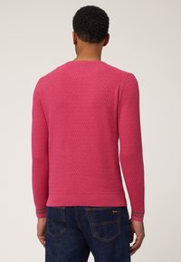 Maglione rosa con texture, colletto rotondo, maniche lunghe, orlo a coste e dettagli a strisce sui polsini. Indossato con jeans scuri.