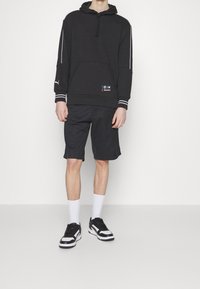 Persona che indossa una felpa nera BMW Motorsport, pantaloni corti neri, calze bianche e sneakers bianco e nere, in piedi contro uno sfondo bianco.
